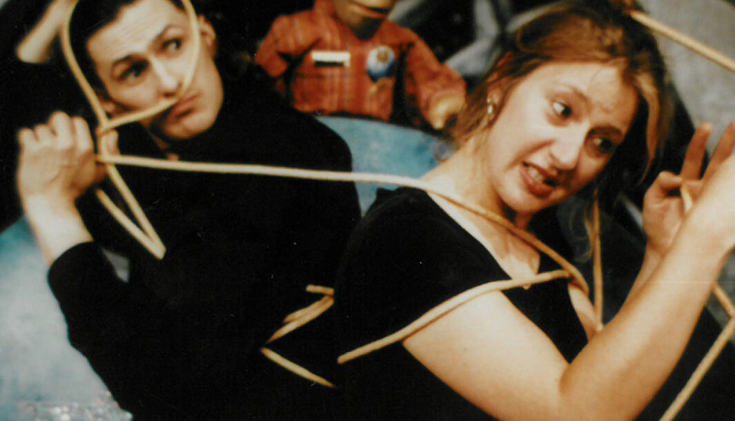 A 'Little Moments' production photo. Two people wearing black are tied in a rope. Behind them is a puppet with red curly hair and wears a red shirt.