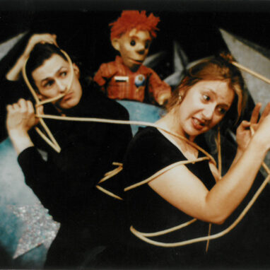 A 'Little Moments' production photo. Two people wearing black are tied in a rope. Behind them is a puppet with red curly hair and wears a red shirt.