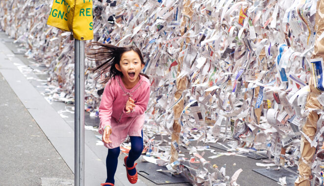 A Sticky Maze production photo. A child in a pink dress runs along a wall of sticky tape covered in hundreds of small pieces of paper. They are smiling widely and their long hair fans out behind them. Photographer: Sarah Walker