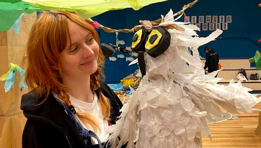 A behind-the-scenes Paper Planet photo. A Polyglot artist holds a large, handmade paper and cardboard owl. She smiles at it. She is in a large school hall, filled with tall, brown cardboard trees.