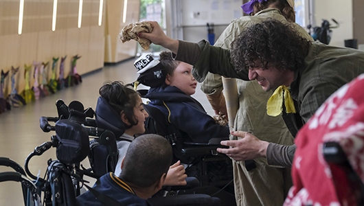 A When the World Turns creative development photo. Polyglot artists in khaki explorer costumes engage with students in wheelchairs. They are in a school corridor. Photo: Suzanne Phoenix