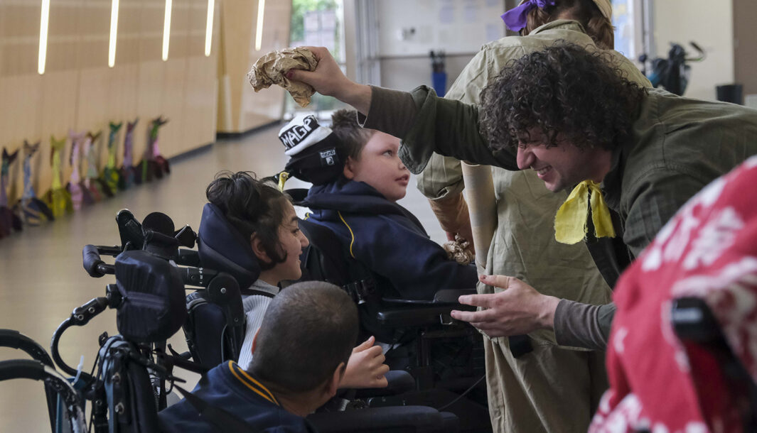 A When the World Turns creative development photo. Polyglot artists in khaki explorer costumes engage with students in wheelchairs. They are in a school corridor. Photo: Suzanne Phoenix