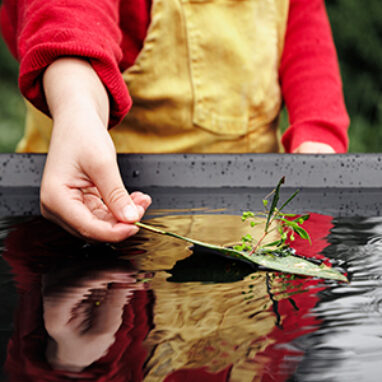A FLOAT promo photo. A child in a red jumper and yellow overalls carefully places a small leaf boat in a pool of rippling water, in which their reflection is visible. Photographer: Darren Gill