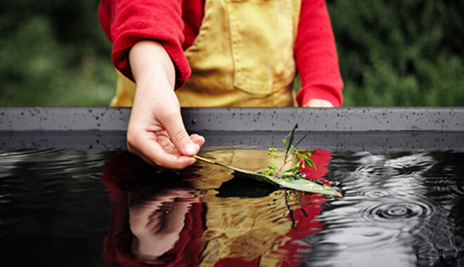 A FLOAT promo photo. A child in a red jumper and yellow overalls carefully places a small leaf boat in a pool of rippling water, in which their reflection is visible. Photographer: Darren Gill