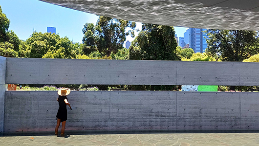 A FLOAT photo. Polyglot artist Afsaneh stands in the MPavilion 10 reflecting pool. She wears black, and a wide-brimmed straw hat, and holds a basket with a long handle up towards the ledge in the back wall. Photographer: Cat Sewell.