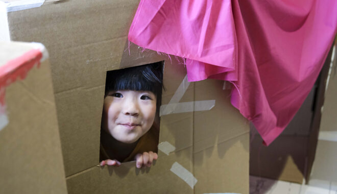 A Cubbyhood photo. A small child peers out of a window in a cardboard box cubby. They smile at the camera. Photographer: Suzanne Phoenix
