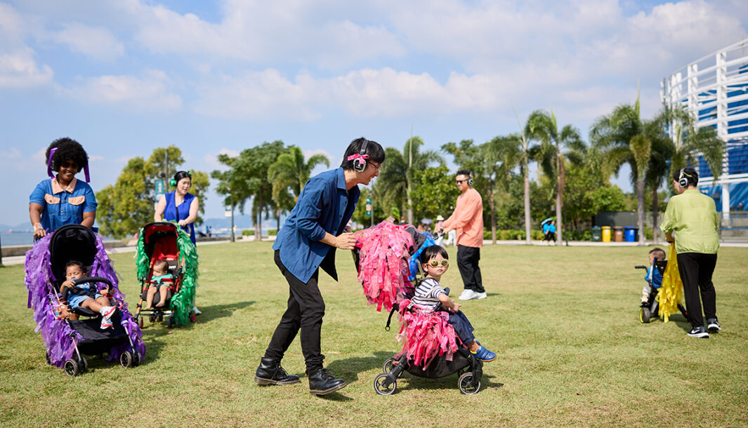 A Pram People (Hong Kong Edition) photo. Adults wearing headphones push their small children in prams decorated with brightly coloured ribbons. They are on a grassy lawn, with a harbour and palm trees in the background. Photo courtesy of Learning and Participation Team, Performing Arts Division, West Kowloon Cultural District.