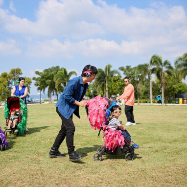 A Pram People (Hong Kong Edition) photo. Adults wearing headphones push their small children in prams decorated with brightly coloured ribbons. They are on a grassy lawn, with a harbour and palm trees in the background. Photo courtesy of Learning and Participation Team, Performing Arts Division, West Kowloon Cultural District.
