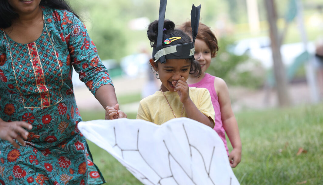 A Bees production photo. Two children, one wearing handmade paper antennae and holding their adult’s hand, joyfully engage with an artist in a Bee costume (only part of their wing is visible). Photographer: Dan Welk, Des Moines Performing Arts