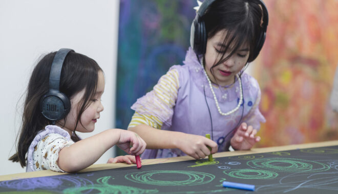 A Sound of Drawing production photo. Two children wearing headphones draw green swirls using oil pastels on a table. The table is lined with black paper. Photo: Theresa Harrison.