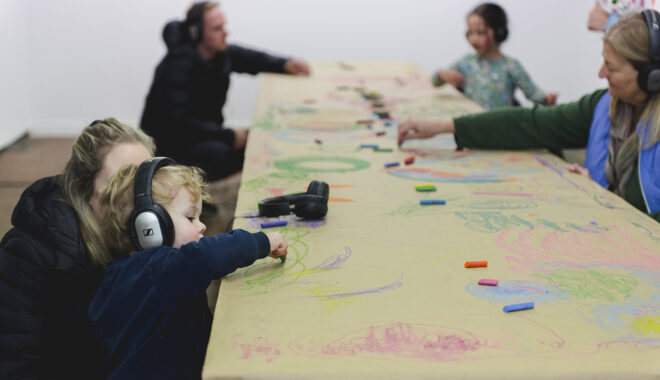 A Sound of Drawing production photo. An adult watches a toddler wearing headphones as they draw on a table lined with brown paper. The table is covered in drawings and pastels. Photo: Theresa Harrison.