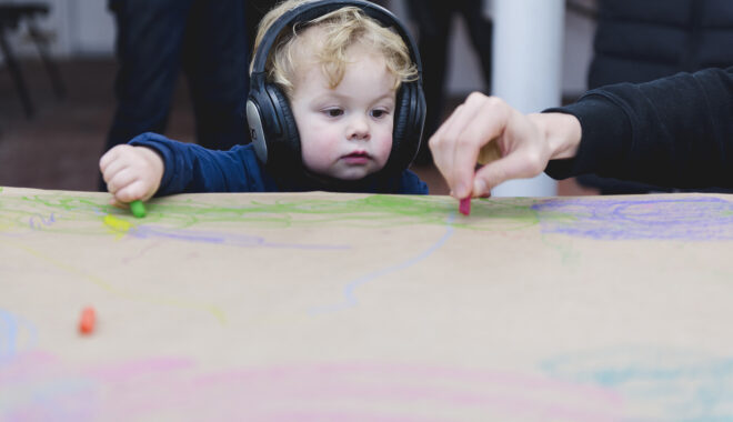 A Sound of Drawing production photo. A toddler watches an adult's hand who holds a pastel as they draw on a table lined with brown paper. The table is covered in drawings. Photo: Theresa Harrison.