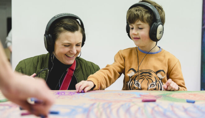 A Sound of Drawing production photo. An adult and a child wearing headphones smile as they draw on a table. The table is covered in drawings and has pastels on it. Photo: Theresa Harrison.