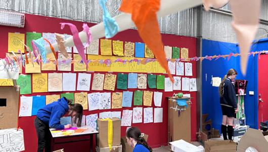 A Wish Street photo. Children create and play with cardboard boxes in a large space, next to a noticeboard covered in drawings and notes on colourful paper. Photo: Sylvie Meltzer & Tamara Rewse