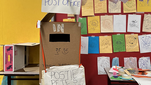 A Wish Street photo. A large cardboard box post office stands in front of a noticeboard covered in drawings and notes on colourful paper. Photo: Sylvie Meltzer & Tamara Rewse