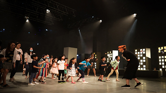 A Come Back Home production photo. A Polyglot artist dressed in black stands in front of a group of children, who are moving towards them.  They are on a large, theatrically lit stage. Photo: Studio ZNKE, Esplanade - Theatres on the Bay, Singapore