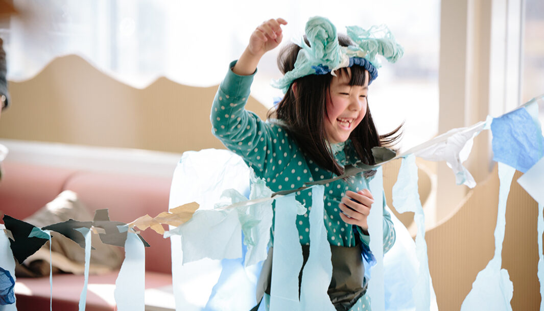 A Paper Planet production photo. A smiling child in a handmade tissue paper costume creates and plays among suspended blue tissue paper creations. Photo: Ai Ueda