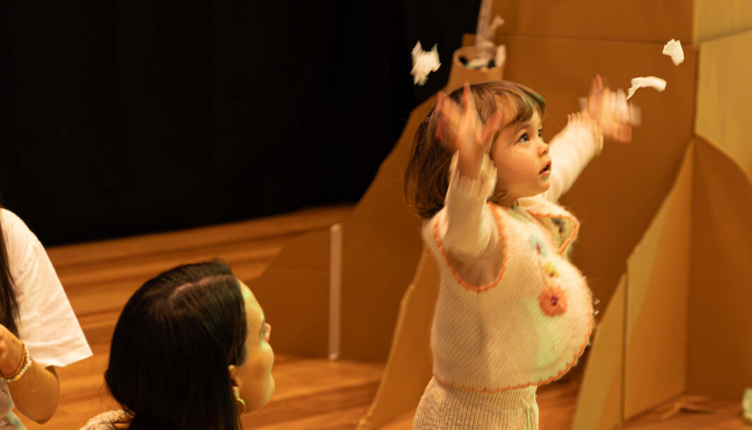 A Paper Planet production photo. A small child throws paper into the air while their adult looks on. They are in a theatrically lit space, filled with tall brown cardboard trees. Photo: Katje Ford, Sydney Opera House.