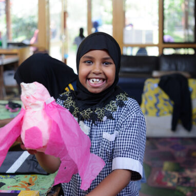 An Us. Here. Now. production photo. A child in a blue and white school uniform and black headscarf smiles at the camera, holding up their pink tissue paper creation. Other children create and play in the background. Photographer: Sarah Walker