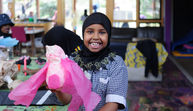 An Us. Here. Now. production photo. A child in a blue and white school uniform and black headscarf smiles at the camera, holding up their pink tissue paper creation. Other children create and play in the background. Photographer: Sarah Walker