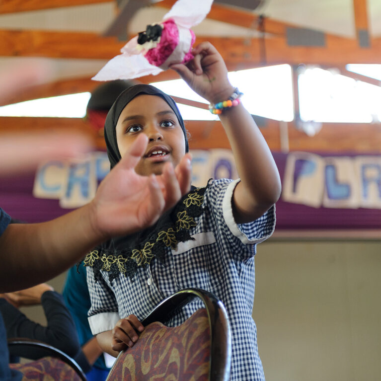 An Us. Here. Now. production photo. A child in a blue and white school uniform and black headscarf flies their tissue paper creation through the air. Photographer: Sarah Walker