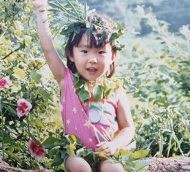 A child sits on a log, wearing a leaf crown and necklace. Their right arm is raised in the air and their fist is clenched.