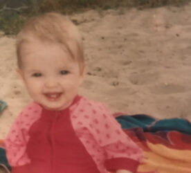 A baby is smiling and sits on sand, wearing a pink bathing suit.