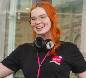 A person wearing glasses smiles. They have orange hair tied up in a ponytail. Wireless headphones rest on their neck. Photo by Theresa Harrison.