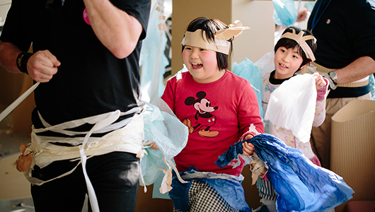 A Paper Planet production photo. Two children, wearing handmade paper costumes, follow a Polyglot artist through tall cardboard trees. Photographer: Ai Ueda