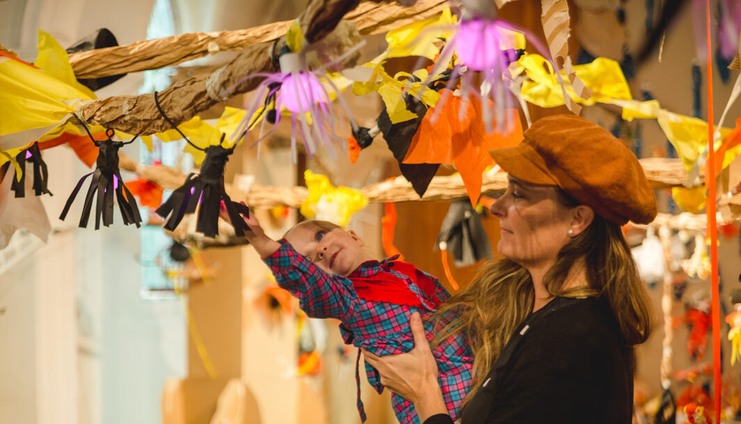A Paper Planet production photo. A parent in a brown hat holds their small child up to a paper vine, adorned with black, yellow and orange paper creatures and plants. The child leans out, reaching towards a creation. Photographer: Theresa Harrison