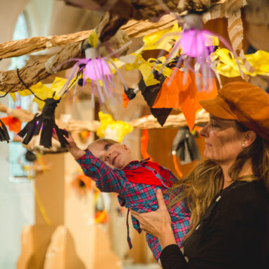 A Paper Planet production photo. A parent in a brown hat holds their small child up to a paper vine, adorned with black, yellow and orange paper creatures and plants. The child leans out, reaching towards a creation. Photographer: Theresa Harrison