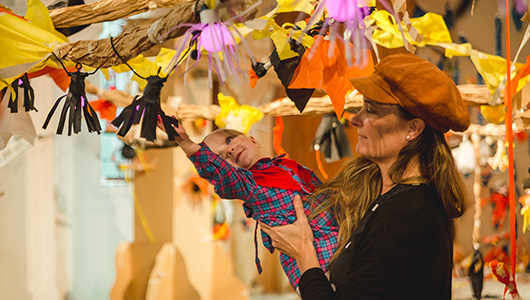 A Paper Planet production photo. A parent in a brown hat holds their small child up to a paper vine, adorned with black, yellow and orange paper creatures and plants. The child leans out, reaching towards a creation. Photographer: Theresa Harrison