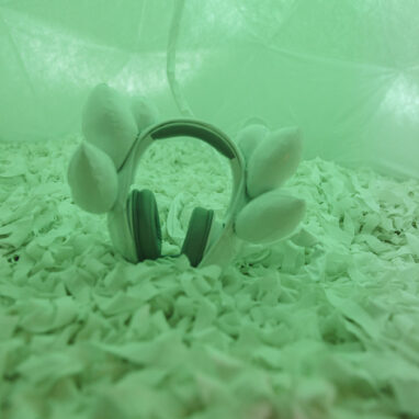 A Voice Lab production photo. A pair of headphones is placed on white carpet, bathed in soft green light. There are white earpieces on the headphones. Photo: Theresa Harrison.