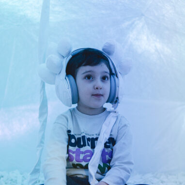 A Voice Lab production photo. A child is seated cross legged in a white dome bathed in warm, white light. They are wearing headphones decorated with white earpieces. Photo: Theresa Harrison.