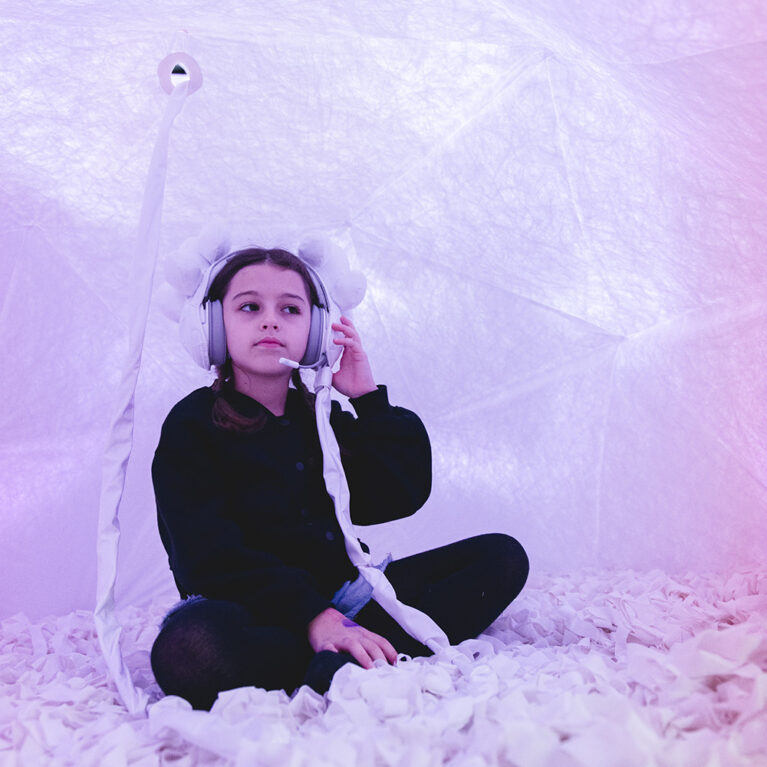 A Voice Lab production photo. A child is seated cross legged in a white dome bathed in warm, pink light. They are wearing headphones decorated with white earpieces. Photo: Theresa Harrison.