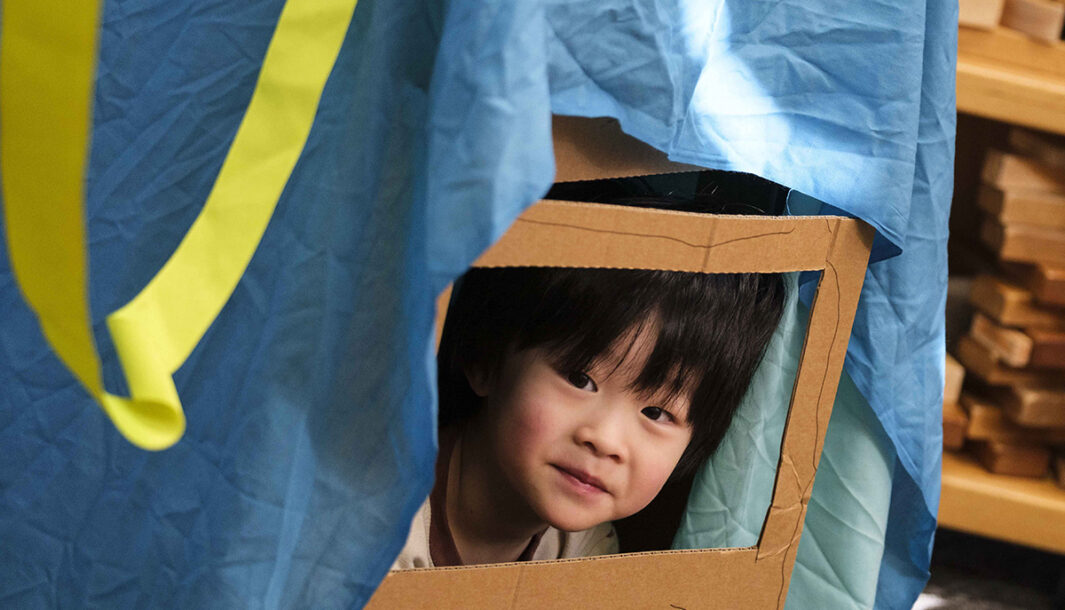 A Cubbyhood workshop photo. A child peers through a cardboard window under a blue piece of cloth. The child has black hair and is smiling. Photo by Suzanne Phoenix.