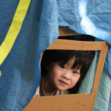 A Cubbyhood workshop photo. A child peers through a cardboard window under a blue piece of cloth. The child has black hair and is smiling. Photo by Suzanne Phoenix.
