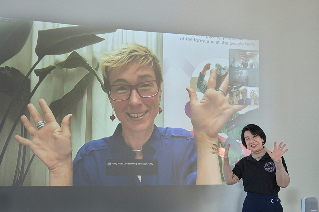 Mikako stands in front of a large screen, filled with a projection of a Zoom meeting with Rainbow. They are both gesturing with their hands and smiling. 