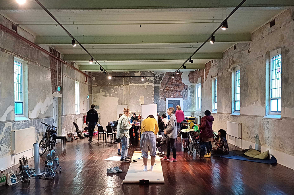 A Catalyst Day photo. Generator artists stand and sit together in a large room, surrounded by materials and chairs. Photo: Cat Sewell