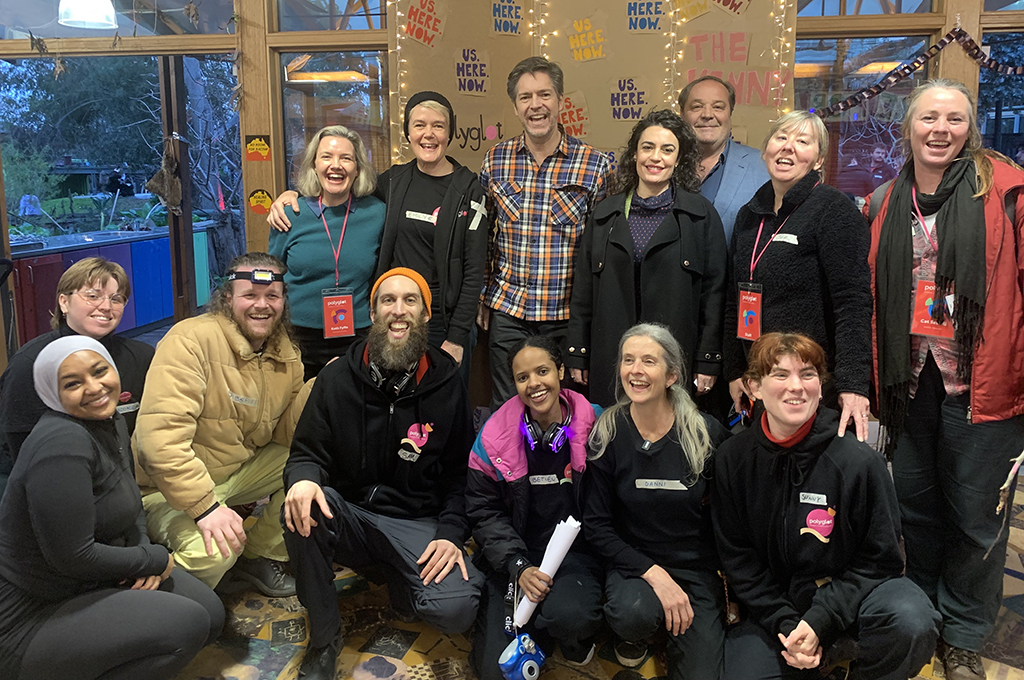 Polyglot artists and staff, The Venny playworkers, and the Lord Mayor of Melbourne, Nicholas Reece, stand together, smiling. They are inside, in front of a handmade cardboard Us. Here. Now. media wall covered in fairy lights.