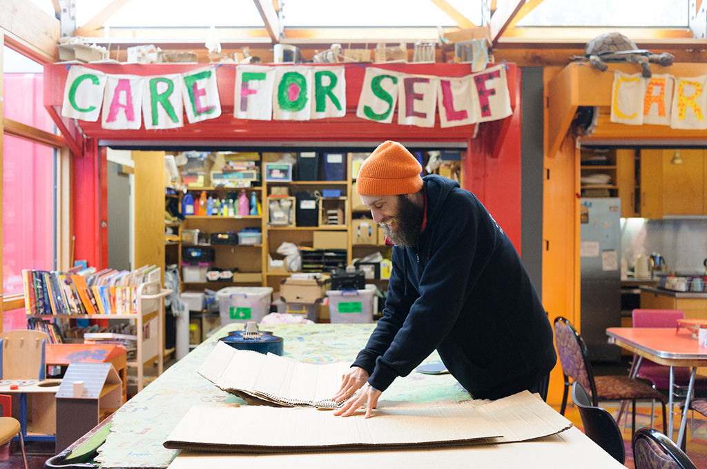 An Us. Here. Now. production photo. A Polyglot artist in a black hoodie and yellow beanie lays materials out on a large table. They are indoors at The Venny, with a handmade sign on the wall behind them: 'Care for self'. Photo: Sarah Walker