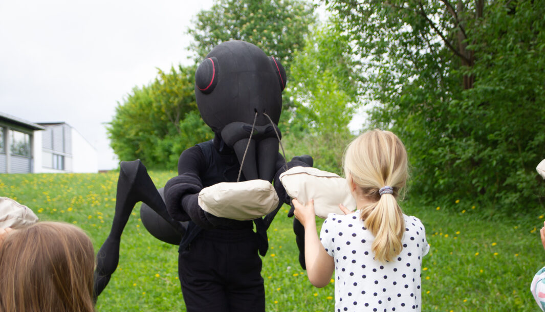 Ants production photo. A Polyglot artist in an elaborate Ants costume passes a child a sack. Photo: Ida M. Halvorsen. 2024.