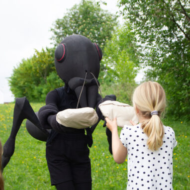 Ants production photo. A Polyglot artist in an elaborate Ants costume passes a child a sack. Photo: Ida M. Halvorsen. 2024.