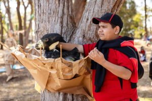 A Totems production photo. A child in a red and black school uniform stands next to a tree, holding their incredible, handmade bird puppet in a paper nest. Photo: Alicia Fox