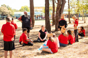 A Totems production photo. Children in red and black school uniforms, and handmade paper costume elements, are gathered outdoors with Polyglot artists Tamara Rewse and Blayne Welsh. They sit and stand under trees. Photo: Alicia Fox