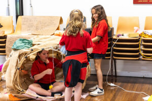 A Totems production photo. Three children wearing red and black school uniforms create and play with paper and bamboo. They are in a room with a wooden floor, next to stacked chairs. Photo: Alicia Fox