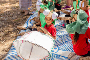 A Totems production photo. Children wearing red and black school uniforms and handmade paper costumes sit together outdoors on a large blue and white rug. One child smiles at the camera. Photo: Alicia Fox