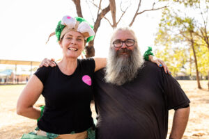 A Totems production photo. Polyglot artists Tamara Rewse and Blayne Welsh stand together, smiling. They wear black t-shirts, and Tamara wears a handmade paper hat. They are outdoors. Photo: Alicia Fox