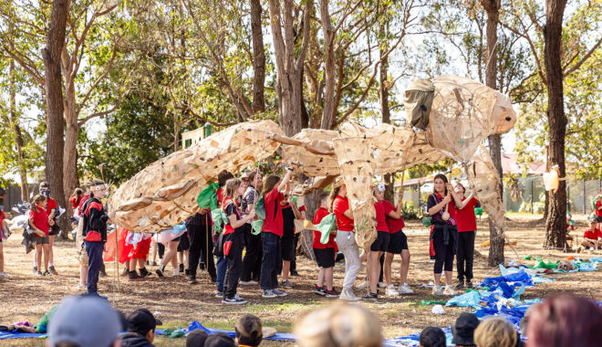 A Totems production photo. Students in red and black uniforms hold up a huge Gurrgiyn / praying mantis made from cane and hessian. They are outdoors, among trees, next to a blue fabric river. Family and friends watch. Photo: Alicia Fox