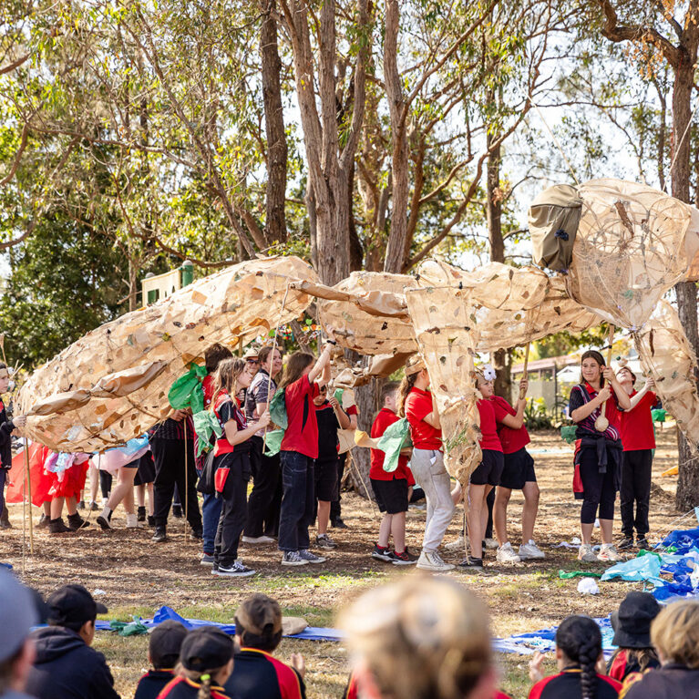 A Totems production photo. Students in red and black uniforms hold up a huge Gurrgiyn / praying mantis made from cane and hessian. They are outdoors, among trees, next to a blue fabric river. Family and friends watch. Photo: Alicia Fox